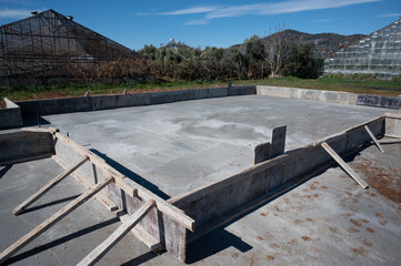 Concrete foundation set within a wooden frame, surrounded by greenhouses and trees, under a clear blue sky. Early stage of building construction