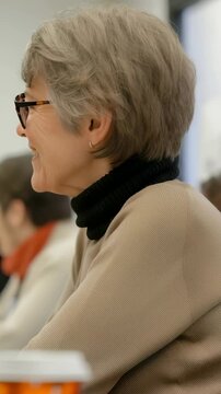 A woman with glasses is participating in a meeting, looking off to the side, with colleagues, data charts and computers on table. Paper cups are scattered across the table surface.