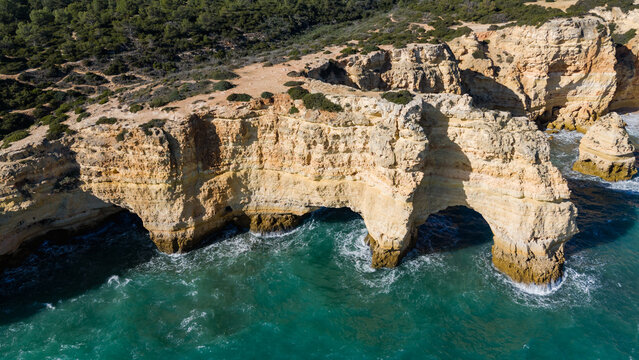 Aerial view of rugged cliffs meet the turquoise sea, arches carved by time stand sentinel against the crashing waves, Estrada da Caramujeira, Lagoa, Faro, Portugal.