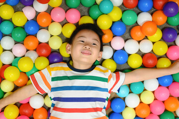Happy asian boy smiling while lying in colorful ball pit, kindergarten playground indoor play area for children, child enjoying fun and childhood happiness.