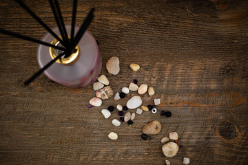 A frosted glass reed diffuser with a gold cap sits on wood, surrounded by pebbles and dark spheres....