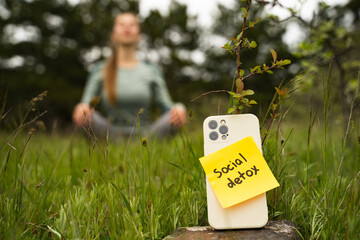 Woman meditating in nature during social detox, blurred background, smartphone with inscription social detox outdoors, stop using digital gadgets, mindfulness and technology break concept