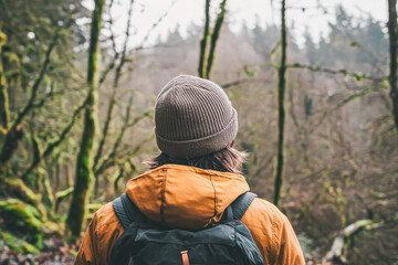 Naklejka premium Man wearing a brown hat and orange jacket is standing in a forest. He is wearing a backpack and has his hands in his pockets. Scene is peaceful and serene, as the man is surrounded by nature