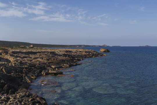 View of a rugged coastline where dark, jagged rocks meet the crystalline turquoise sea under a vast, pale blue sky, Dipkarpaz, Northern Cyprus, Cipro.