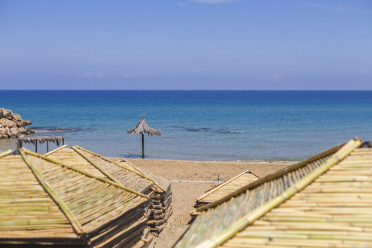 View of golden sands meeting turquoise waters under a clear blue sky, framed by rustic bamboo umbrellas and a solitary straw parasol, Dipkarpaz, Northern Cyprus, Cipro.