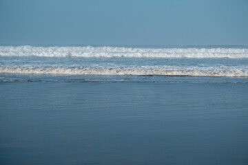 Obraz premium Multiple layers of ocean waves rolling towards the shore under a clear sky, showcasing the dynamic power and beauty of the sea.