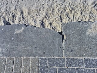 Close up of the edges of chipped concrete pavers on the footpath overhanging sand at the beach. The footprints of seagulls can be seen in the sand.