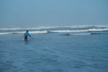 Naklejka premium Yogyakarta, Indonesia-July 3, 2022: A lone fisherman pulling a net in the shallow ocean waters at the beach, under a vast sky, depicting traditional livelihood.