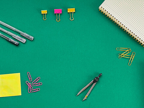 A textured green background with various stationery items. Includes pens, paper clips, binder clips, a yellow sticky