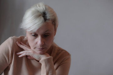 Thoughtful mature woman in beige sweater sitting indoors, resting her chin on her hands, looking...