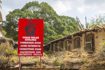 View of a red forbidden zone sign with a soldier silhouette amidst crumbling buildings and overgrown foliage, Varosha, Famagusta, Cyprus.