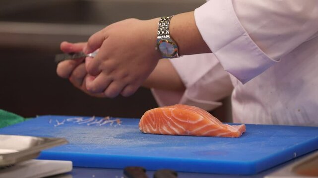Chef's hands meticulously remove pin bones from fresh fish fillets using tweezers. This precise technique on a blue cutting board ensures high-quality, boneless fish for cooking.