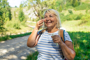 A cheerful senior woman standing on a scenic forest trail applies face cream or sunscreen while...