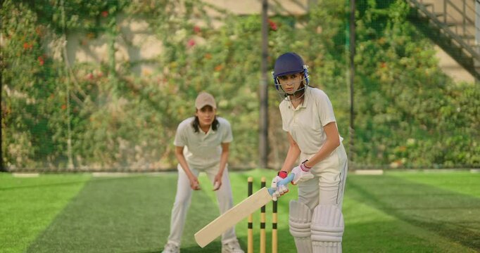 Indian gen z young adult athlete sports woman girl standing at outdoor match play ground wear gloves white uniform protective helmet Asian cricket player female lady hold bat hitting ball do practice
