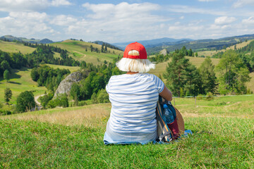 A senior woman with a red cap and striped shirt sits peacefully on a grassy hillside, gazing out at...