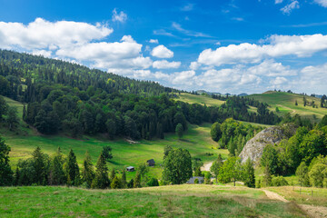 A breathtaking panoramic view of a lush green valley surrounded by forested hills and distant mountains under a bright blue sky with scattered clouds. 