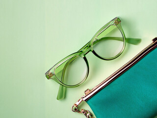 Flat lay with a chic pair of glasses with a transparent green frame and a green clurch bag on a plae green background with copy space to the left.