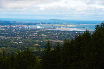 Expansive panoramic view of Dublin, featuring urban sprawl, industrial structures with chimneys, and a wide bay under a cloudy sky. Evergreen trees frame the foreground