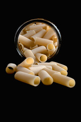 Uncooked rigatoni pasta tubes overflowing and spilling out from a clear glass jar onto a black,...