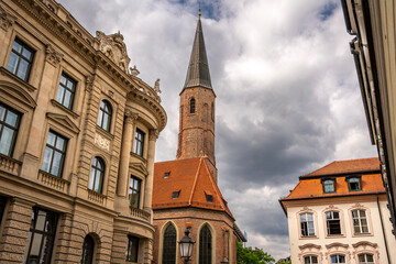 Historic St. Jakobs Church in Munich Near Sendlinger Tor: Elegant Gothic Architecture with Tall Steeple and Red Roof Surrounded by Classic Bavarian Buildings under Dramatic Cloudy Sky