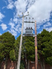 High voltage transformer sat on a wooden Electricity Pylon in front of trees against a blue cloudy...