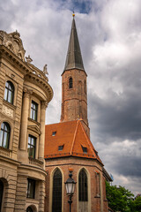 Historic St. Jakobs Church in Munich Near Sendlinger Tor: Elegant Gothic Architecture with Tall Steeple and Red Roof Surrounded by Classic Bavarian Buildings under Dramatic Cloudy Sky