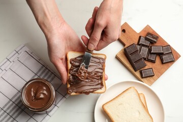 Woman spreading tasty chocolate butter onto bread at white table, top view