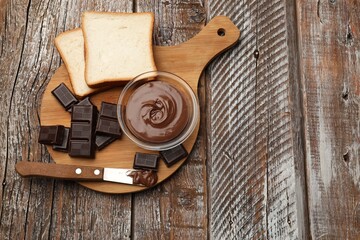 Tasty chocolate butter in glass bowl, slices of bread and knife on color wooden table, top view....