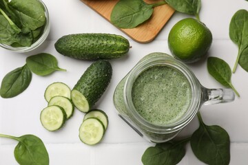 Tasty smoothie in mason jar, spinach leaves, lime and cucumber on white tiled table, flat lay