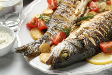 Tasty roasted seabass fish served with potatoes, tomatoes and lemon on white marble table, closeup....
