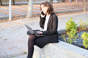 Executive woman sitting with laptop on lap talking on smartphone at sunset