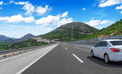 A white car drives along a smooth mountain highway under a bright blue sky with scattered clouds,...