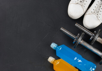 Flat lay image featuring a pair of white sport shoes, two metal dumbbells, and bottles of blue and orange energy drinks on a dark textured background. The composition highlights essential items for fi