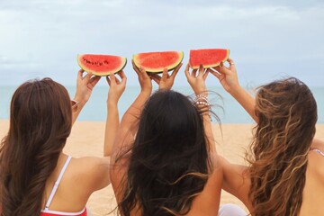 Three young Asian women having fun on a tropical beach, kneeling on the sand and holding watermelon slices on their heads.