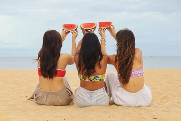 Three young Asian women having fun on a tropical beach, kneeling on the sand and holding watermelon...