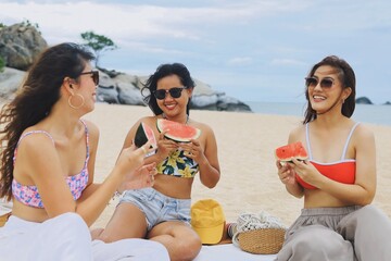 Three happy young Asian women enjoying pizza and drinks during a beach picnic on a sunny summer...