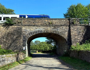 A Victorian stone bridge spans a dirt path beneath a passing train, framed by lush foliage and open sky, with sunlight sharpening the texture in Norwood Green, Halifax, UK.