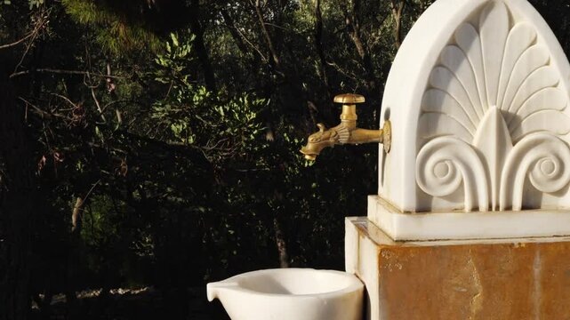 Detailed close-up of a traditional Greek marble water fountain near the Odeon of Herodes Atticus in Athens. Shows ornate brass spout, carved shell, and floral designs. No people visible.