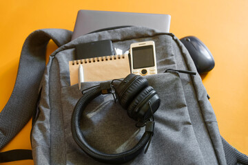 Top‑view of audio essentials including cushioned headphones, mobile phone, notebook and power bank on a grey college backpack against a yellow backdrop. High resolution