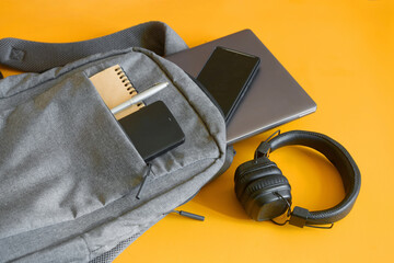 Top‑view of a student or traveler’s backpack filled with modern gadgets—laptop, phone, power bank, notebook and pen—beside cushioned headphones on a yellow backdrop. High resolution