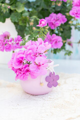 Pink geranium flowers arranged in a decorative purple vase with wooden flower embellishments, placed on a white doily, with additional geraniums visible in soft focus in the background,vertical