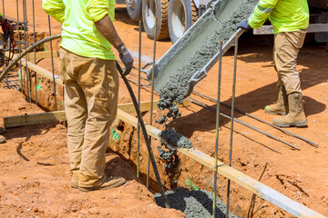Worker in bright clothing pour concrete into foundation trench at construction site under works day...