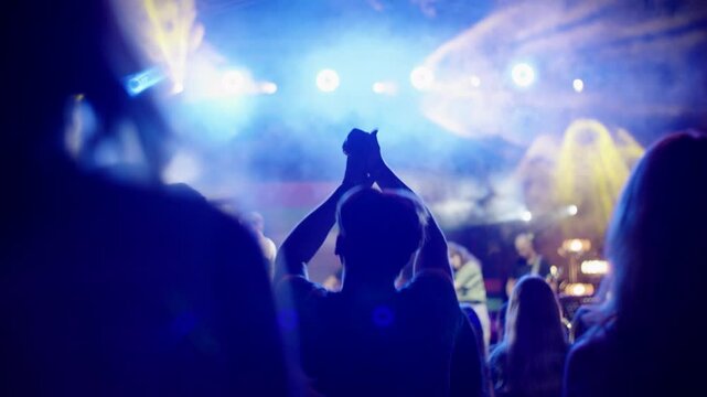 Energetic crowd enjoying live music at a nighttime open-air concert. Fans are jumping and clapping to the rhythm under stage lights. Backside view captures atmosphere and movement.