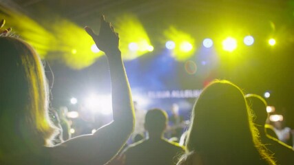 Person raises hand at outdoor concert or worship event, with colorful stage lighting and blurred background. Ideal for music, celebration, church or festival atmosphere footage. Backside medium shot