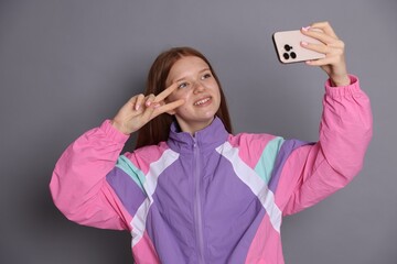 Smiling teenage girl taking selfie by smartphone and showing peace sign on grey background