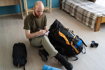 Traveler bearded man writing checklist for hiking trip in mountains sitting on floor at home