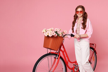 Beautiful teenage girl in sunglasses and bicycle with basket of flowers against pink background....