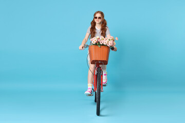 Beautiful teenage girl in sunglasses riding bicycle with basket of flowers against light blue background