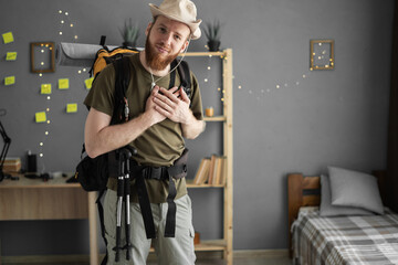 Young bearded man traveler in hat prepare for hiking vacation at home putting hands on chest...