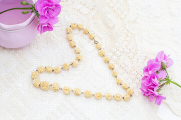Vintage ivory necklace with rose-shaped beads lies on a white lace tablecloth in the shape of a heart, next to purple geranium flowers and a lilac vase, creating a delicate and romantic composition,